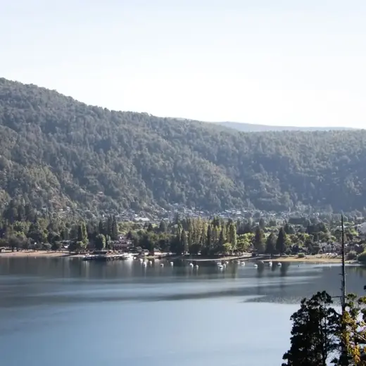 Costa de un lago con cabañas entre los árboles y montañas al fondo