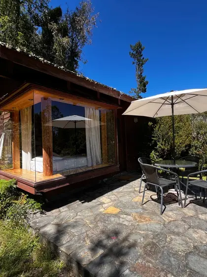 Cabaña de madera con un gran ventanal, sombrilla y terraza de piedra