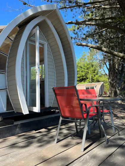 Tiny house de techo curvo con terraza de madera y sillas rojas entre pinos